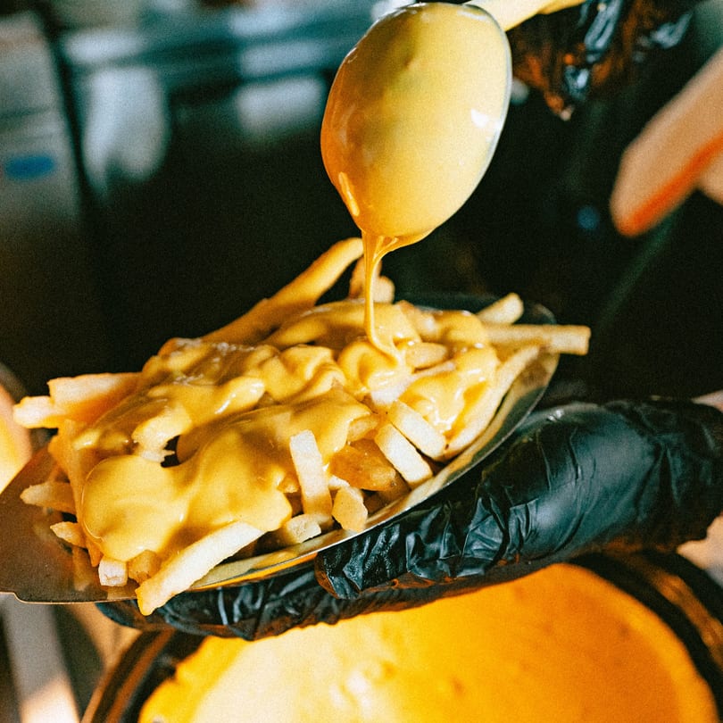 A gloved hand holds a tray of french fries as cheese sauce is poured over them.