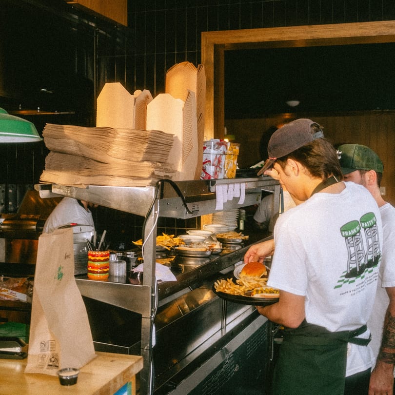 Two cooks preparing burgers and fries in a busy kitchen with takeout containers stacked.