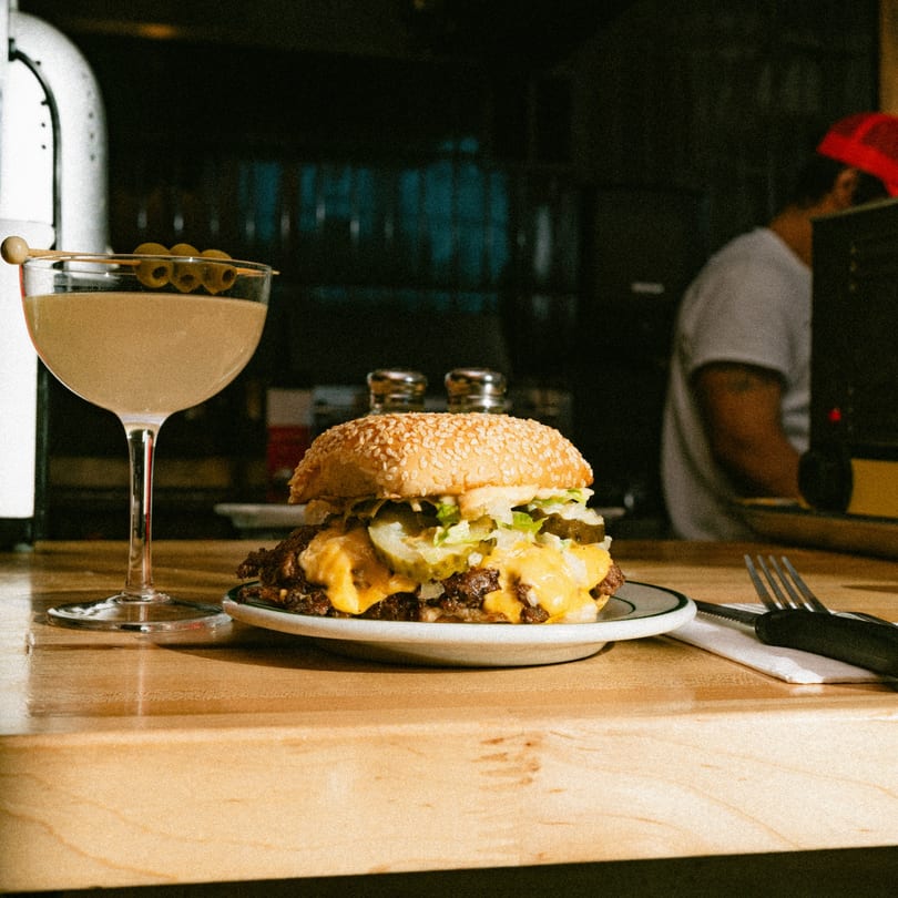 A large cheeseburger and a martini with olives on a wooden bar counter.