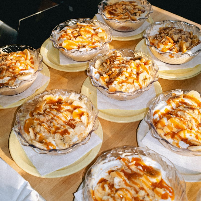 Several glass bowls of banana pudding with caramel drizzle and cookie crumbles on yellow plates.