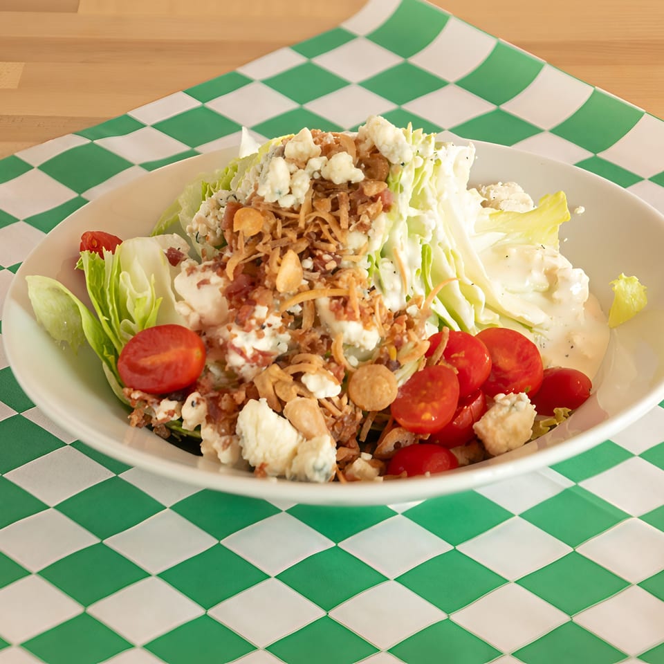 Wedge salad with blue cheese, cherry tomatoes, and crispy fried onions on a green and white checkered tablecloth.