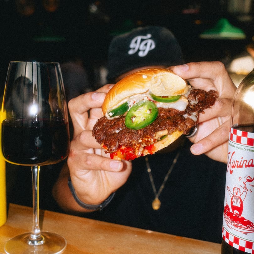 Hands holding a juicy burger with jalapeños and red peppers, next to a glass of red wine.