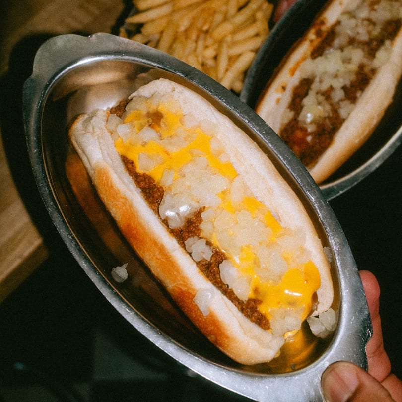 A chili cheese hot dog with diced onions in a metal tray, with fries in the background.